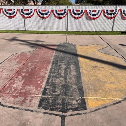 Basketball court with large abstract design from KA House Painting Project by Piper Family, showcasing red, black, and yellow weathered paint, festive bunting, and community spirit.