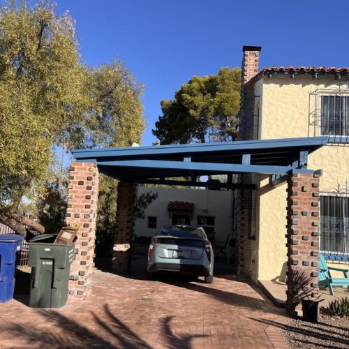 Charming Tucson residential scene featuring a carport with brick pillars and a blue roof, a sandy yellow Spanish-style house, bins on a brick driveway, lush tree, and drought-tolerant plants.
