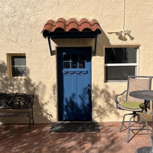 A serene Southwest patio features a warm stucco wall with a vibrant blue door and terracotta awning, surrounded by rustic bricks and shaded greenery, evoking a peaceful Tucson charm.