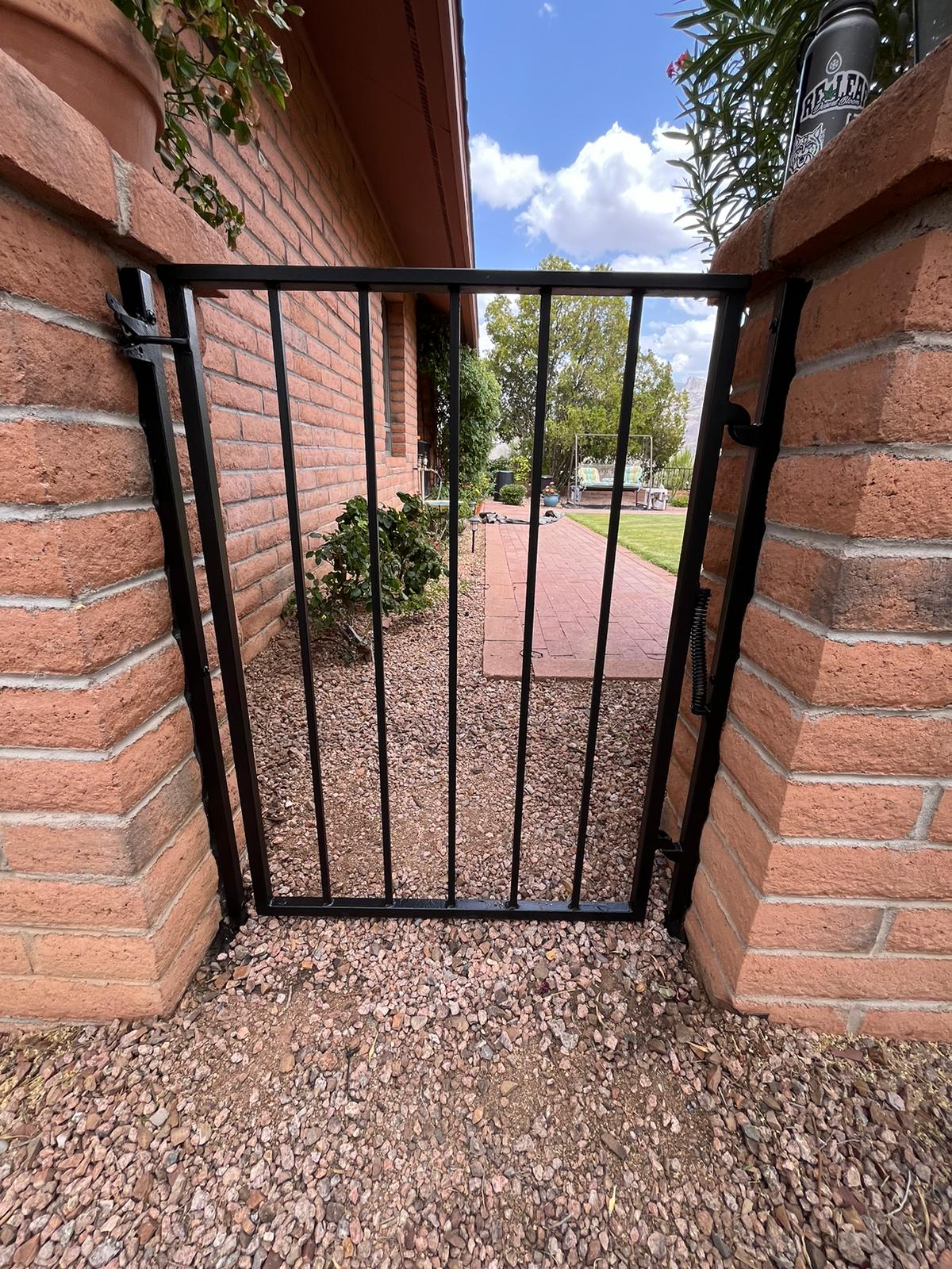 Gate and Fence Painting in Tucson featuring a sleek black metal gate between brick pillars with a garden view and blue sky backdrop.